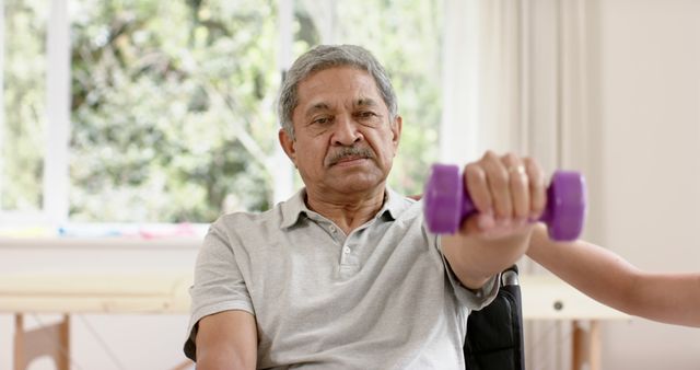 Elderly man lifting dumbbells in a physical therapy session. He seems to be focused on his exercise routine, emphasizing the importance of maintaining physical fitness and strength in senior years. Indoor setting with natural light coming through windows. Ideal for topics related to senior fitness, rehabilitation, healthy aging, physical therapy, and wellness programs for the elderly.