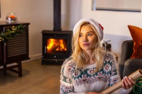 Blonde woman wearing santa hat wrapping gifts by wood stove in cozy holiday living room