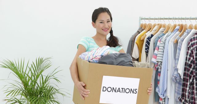 Pretty asian brunette holding donation box full of clothes smiling at camera