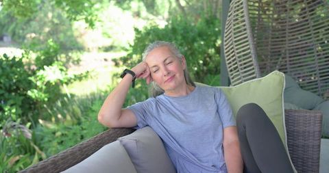 Senior woman relaxing on wicker patio sofa with wrist tracker enjoying peaceful garden