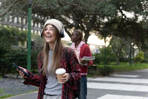 Smiling young woman listening to music walking campus path holding coffee and phone
