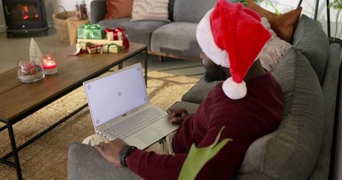 African American man sitting on sofa wearing Santa hat using laptop by fireplace and gifts
