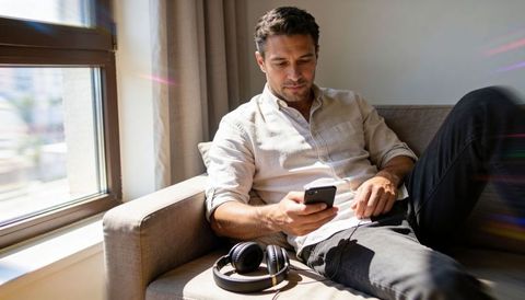 Relaxing man using smartphone on sunlit sofa by window with headphones, casual shirt