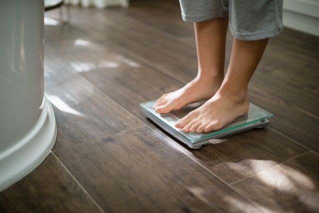 Boy standing on weight scale in a home environment with hardwood flooring. Ideal for use in articles or advertisements related to health, fitness, weight management, and healthy lifestyle tips. Can also be used in blogs or social media posts about personal health journeys and self-care routines.