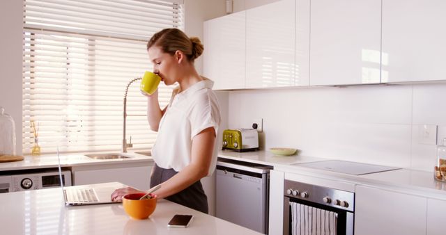 Woman using laptop while having coffee in kitchen at home 4k