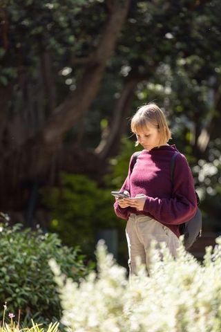 Female student texting on smartphone in sunny campus garden with backpack and greenery