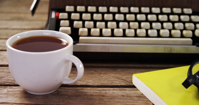 White coffee cup filled with coffee placed next to a vintage typewriter on a wooden desk. Yellow notebook and glasses are present, adding to the nostalgic, old-fashioned workplace atmosphere. Perfect for illustrating concepts related to retro workspaces, nostalgia, office settings, literary themes, and productivity.