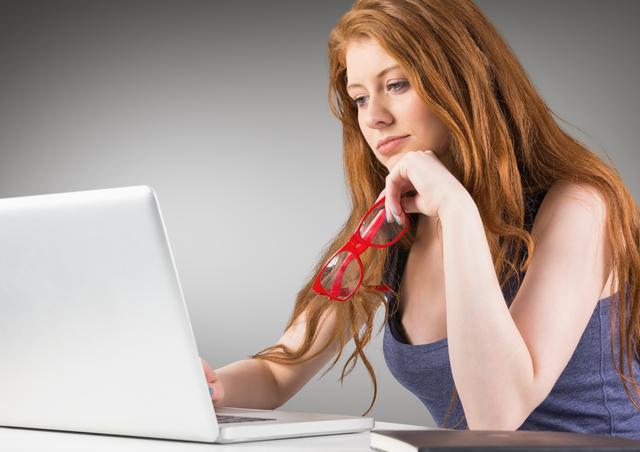 This image depicts a woman with red hair using a laptop, holding red glasses in her hand. She appears focused and is likely working or studying. This image can be used for articles or advertisements related to technology, remote work, education, or modern office environments.