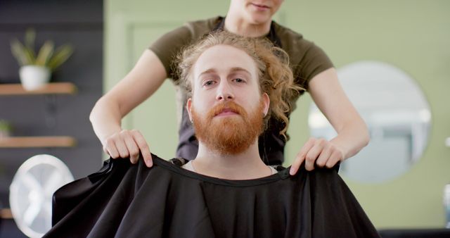 This stock photo captures a redheaded man being prepared for a haircut by a barber. The focus is on the man’s confident expression as the barber readies him by adjusting the cape. Suitable for use in content related to men's grooming, barber services, and hairdressing promotions. It can also be used for blogs or commercials focusing on beard and hair care.