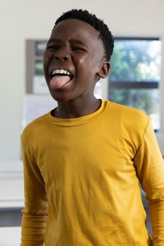 Playful african american boy sticking out tongue in mustard shirt classroom portrait