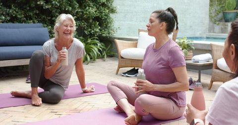 Diverse women practicing yoga, laughing and hydrating on sunlit patio near pool