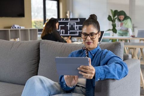 Young professional using tablet on sofa in modern coworking lounge with collaborative team