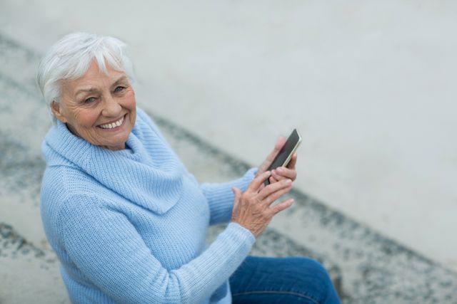 Senior woman sitting on steps, using mobile phone and smiling. Ideal for themes related to elderly people embracing technology, modern communication, active retirement, and outdoor leisure activities.