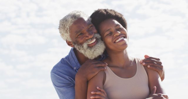 Senior man hugging smiling woman outdoors with clear sky in background. Perfect for use in family and relationship-themed campaigns, promotional materials for family-oriented products or services, and websites focused on bonding and love.