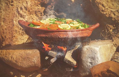 Rustic clay pot cooking over open fire with lemon slices, tomatoes and fresh herbs