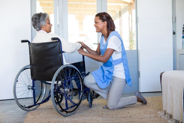 Caregiver kneeling and interacting with senior woman in wheelchair, providing support and assistance in a home setting. Ideal for use in healthcare, elderly care, and home care service promotions, as well as articles on senior living and medical support.