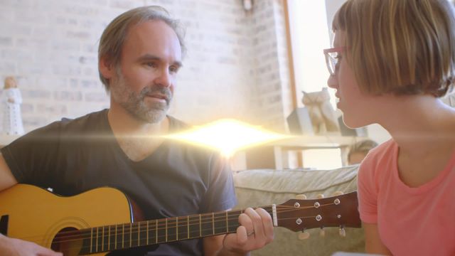 Father is seen playing an acoustic guitar while singing along with his daughter, suggesting a bonding moment at home. This high-quality image captures the essence of family time, music enjoyment, and parent-child connection, making it ideal for articles, blogs, advertisements, and promotional materials that focus on family life, parenthood, and music-related content.