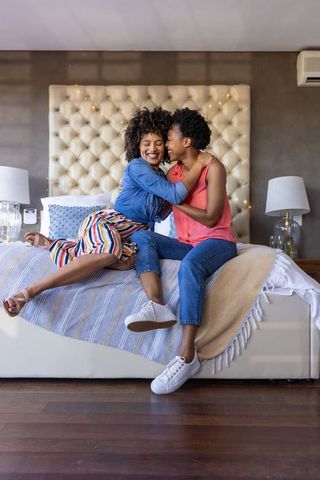 Black women couple embracing and laughing on bed with tufted headboard and string lights