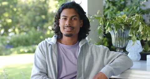 African american man relaxing on porch railing wearing hoodie amid sunlit garden greenery