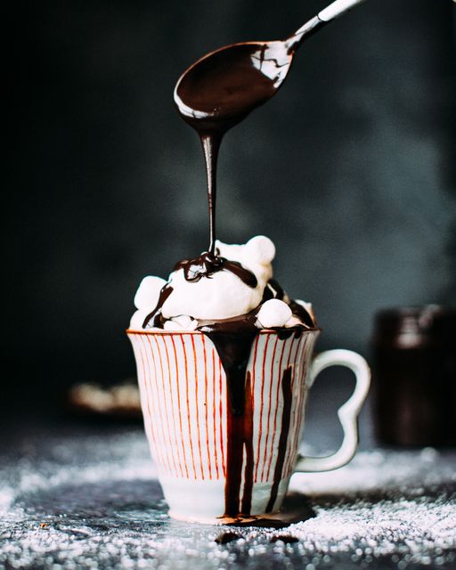 This image shows a handmade ceramic mug filled with hot chocolate, crowned with whipped cream and marshmallows while being drizzled with rich chocolate syrup. Perfect for use in food blogs, dessert recipe sites, café menu designs, or winter-themed advertisements showcasing cozy and indulgent treats.