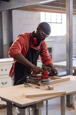 Carpenter using electric sander smoothing wooden plank on workshop workbench