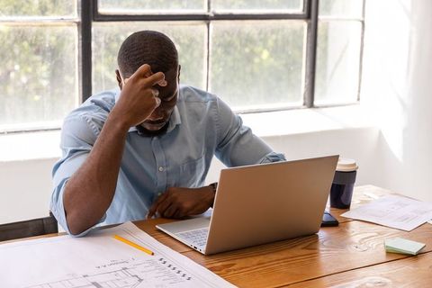 Architect Focused on Blueprint Design Using Laptop in Sunlit Office