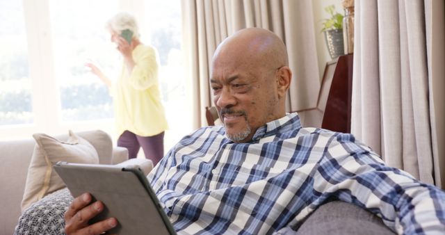 Senior Man Using Tablet at Home While Woman in Background is on Phone Call - Download Free Stock Images Pikwizard.com