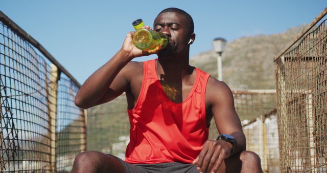 Athletic man hydrating after an intensive outdoor workout, wearing red sports attire and holding a water bottle. Ideal for promoting fitness products, hydration solutions, sports equipment, and healthy lifestyle campaigns. Visually appealing representation of post-exercise recovery and maintaining proper hydration.
