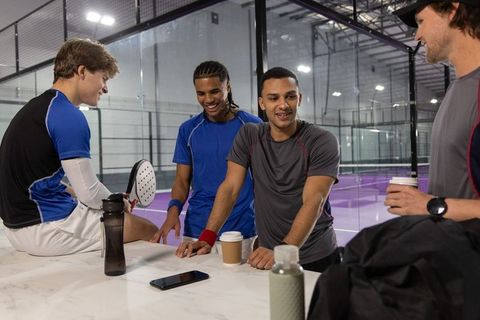 Padel players chatting at indoor glass court counter with paddles, coffee and gear