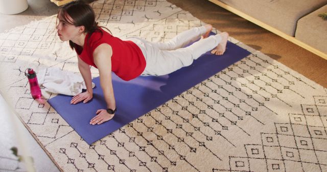 Young woman dressed in a red top and white leggings is practicing a yoga pose on a blue mat in her living room. The rug adds a cozy touch to the surroundings. Perfect visual for articles and posts about home workout tips, yoga routines, indoor physical exercise, healthy living, and self-care.