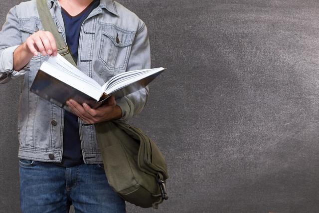 Midsection of young man reading open book while standing against a gray background, wearing denim jacket and carrying a bag. Perfect for use in educational materials, articles about casual fashion, or imagery for learning enthusiasts.