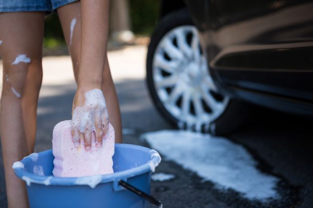 Photo depicts a teenage girl washing a car using a soapy sponge and a bucket of water. Ideal for use in articles or advertisements related to car care, summer activities, hands-on chores, teen lifestyle, or DIY tasks. The bright and sunny environment suggests leisure and outdoor enjoyment.