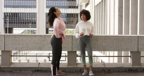 Diverse women in hoodies and leggings chatting, stretching and leaning on urban terrace railing