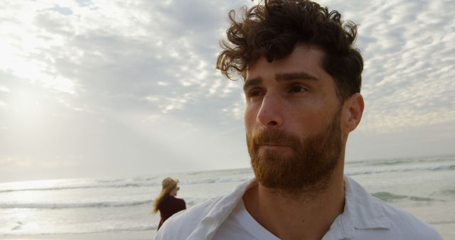 Outdoors scene with a man standing on a beach near the sea, staring into the distance with a thoughtful expression. The sky is cloudy and overcast, with the sunlight diffused through the clouds. Ideal for use in promotional materials for travel and leisure, inspirational content, mental health and wellness campaigns, or as a visual for themes of contemplation and calmness.