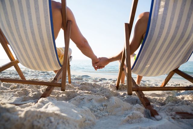 Couple holding hands while relaxing on lounge chairs at beach during sunny day