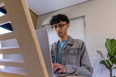 Asian man standing at slatted home workstation working on laptop in denim jacket and glasses