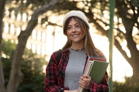 Smiling female student holding notebooks on college campus at golden hour backlit portrait