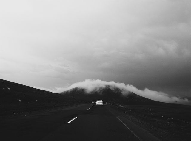 The photograph captures a solitary white car traveling on an empty road leading towards a fog-enshrouded hill under an overcast sky. The absence of color emphasizes the dramatic, moody atmosphere, conveying a sense of solitude and introspection. Ideal for projects centered on travel, introspection, nature, or isolated landscapes, and suitable for use in blog posts, websites, and print media that require evocative, atmospheric imagery.