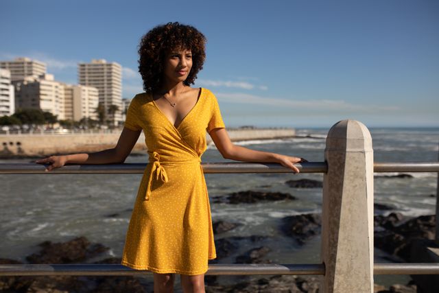 Front view of a biracial woman wearing a yellow dress leaning against a balustrade on a sunny day by the sea