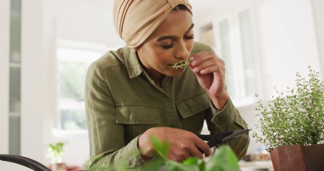 A woman wearing a turban enjoys the aroma of fresh herbs in her kitchen. Ideal for content on indoor gardening, healthy living, culinary activities, and lifestyle blogs. The image portrays a sense of calm and appreciation for natural elements indoors.