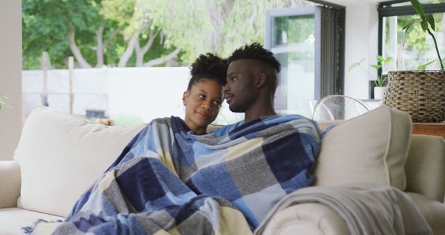 Couple sitting on couch wrapped in blankets, showing affectionate bond. Great for themes of relationships, home comfort, love, and lifestyle promotions.
