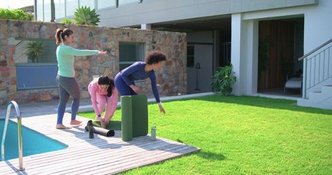 Three women preparing outdoor yoga on backyard deck with mats, foam roller, water bottle