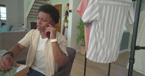 African american man talking on phone while unpacking clothes from box near clothing rack