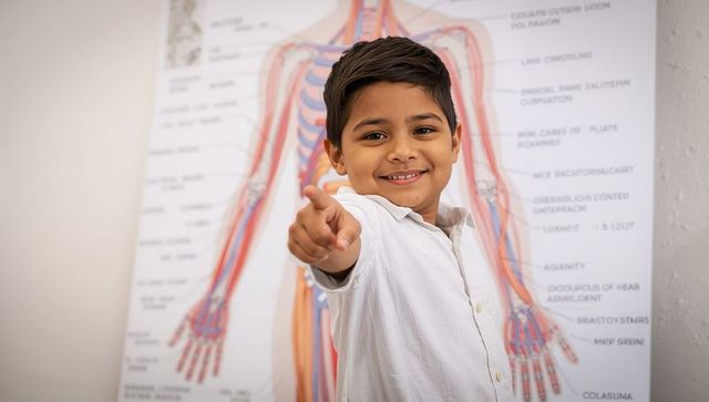 Smiling schoolboy pointing at camera in science classroom with colorful anatomy chart