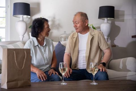Senior asian couple laughing and talking over wine on cozy living room sofa