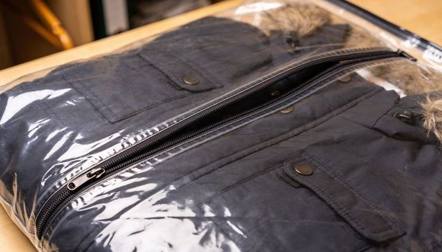 Displaying folded dark parka sealed in clear plastic packaging with visible zipper pull