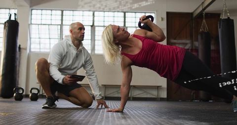 Performing kettlebell side plank row while coach tracks progress on tablet