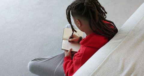 Young man journaling on carpet in red hoodie sitting cross-legged, cozy minimalist room