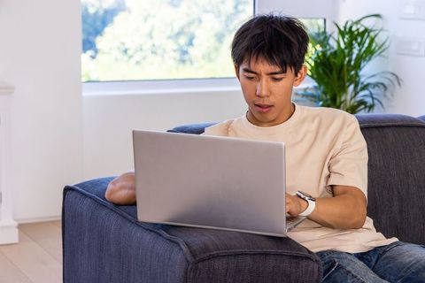 Asian man working on laptop at home on sofa with smartwatch in minimalist living room