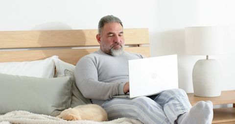 Senior man working on laptop in bed with sleeping dog, cozy loungewear morning